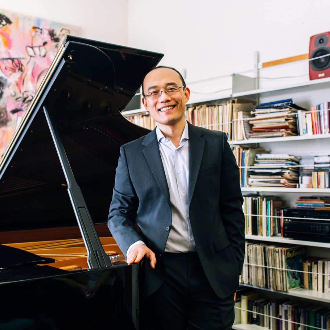 Pianist Dr Jian Liu stands, smiling, next to his piano set against shelves full of manuscripts and music in his office